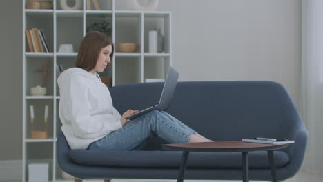 Low-angle-view-of-a-woman-sitting-on-a-sofa-looking-at-her-laptop.-Freelance-Career.-Cheerful-Lady-Working-On-Laptop-Computer-Sitting-On-Sofa-At-Home.